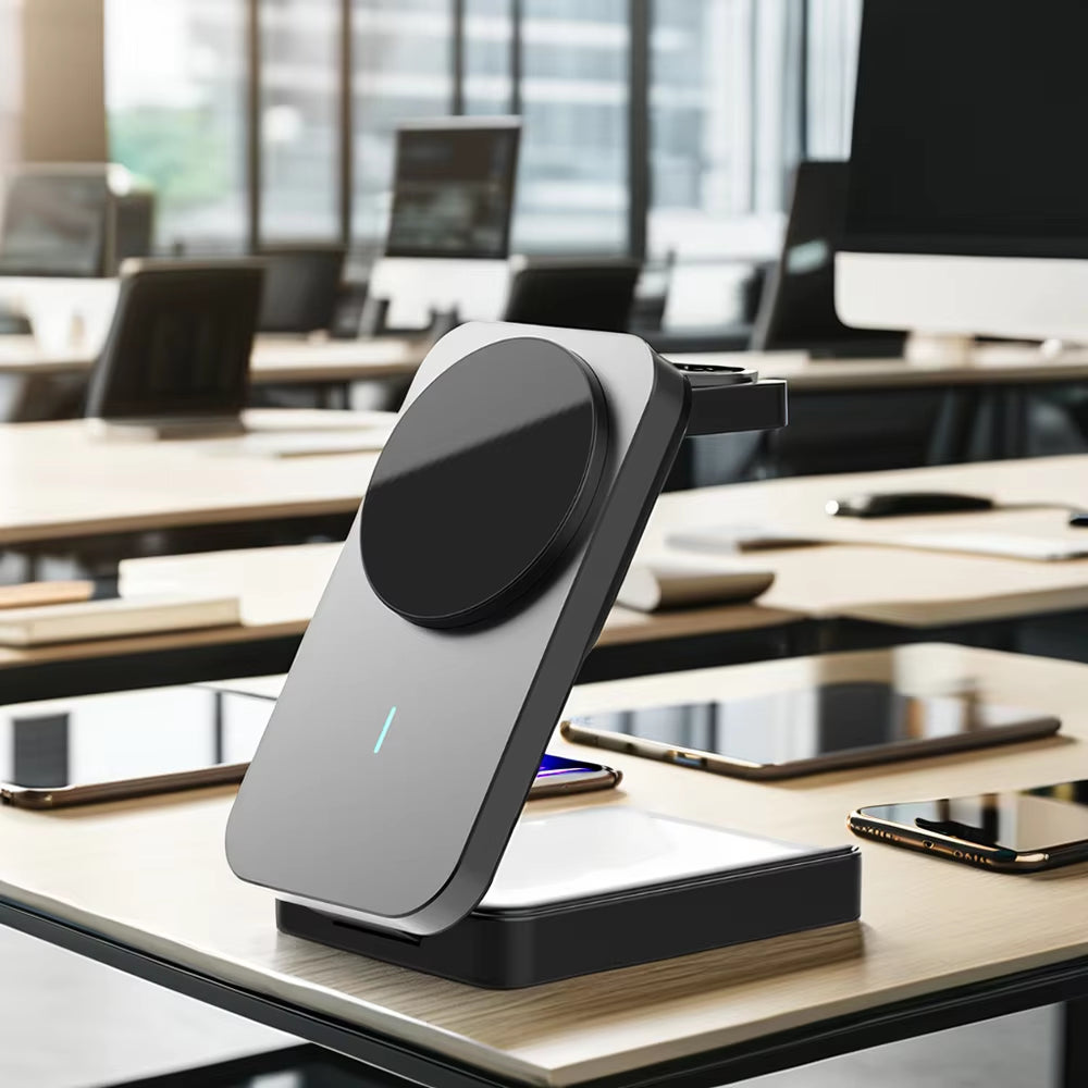 A modern wireless charging stand with a circular magnetic pad, shown on a light wood office desk with smartphones and tablets nearby, and blurred computer monitors in the background.